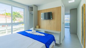 A modern bedroom with a bed, a TV mounted on a wooden panel, and a view of the outside through large windows. - Ocean Blue #01 - Apt With Pool by Carpediem (Ipojuca)