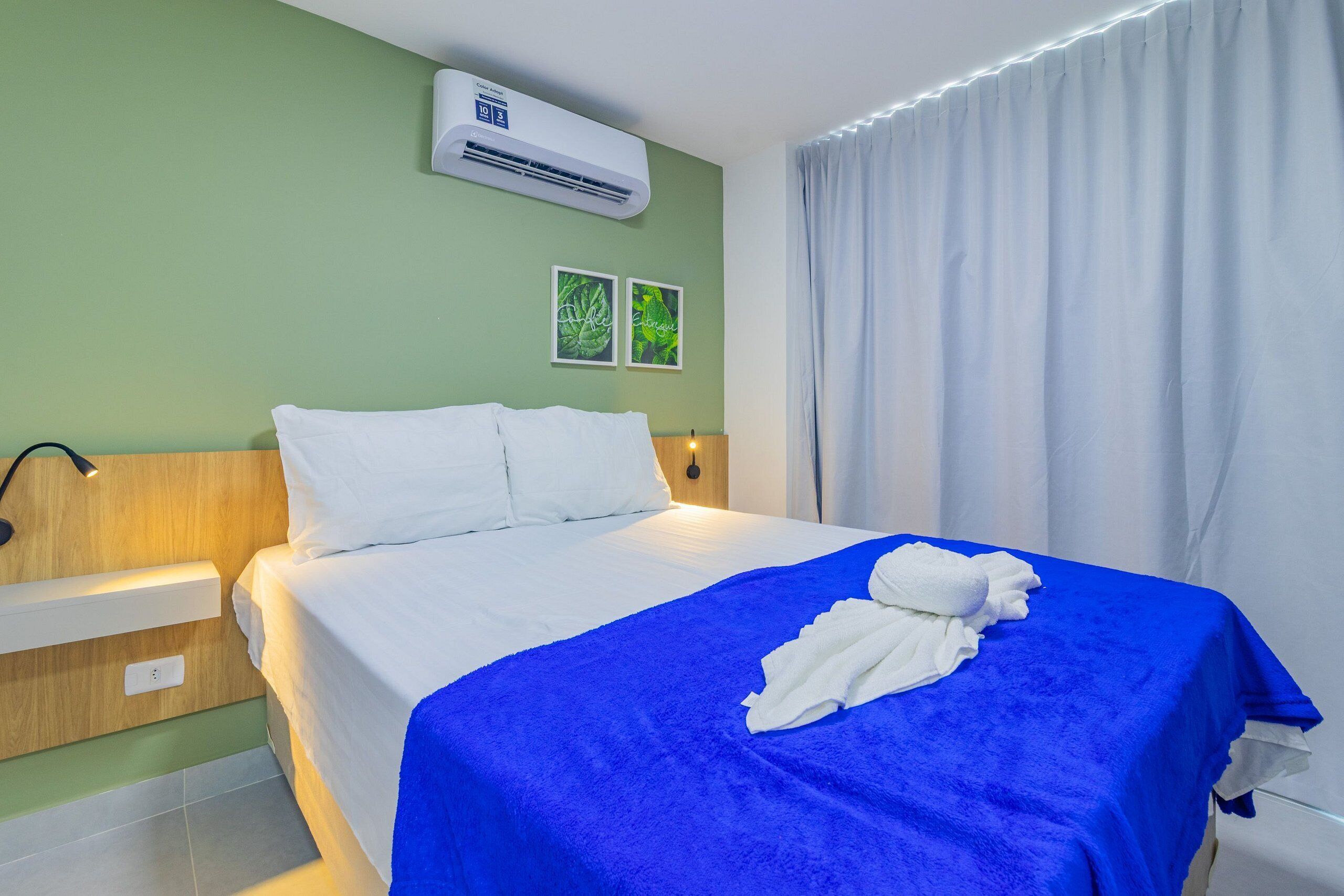 A hotel room with a bed, a blue blanket, white pillows, a wall-mounted air conditioner, and two framed pictures.