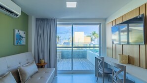 A modern hotel room with a sofa, a dining area with chairs, a TV, and a balcony with a view of buildings and palm trees. - Ocean Blue #01 - Apt With Pool by Carpediem (Ipojuca)