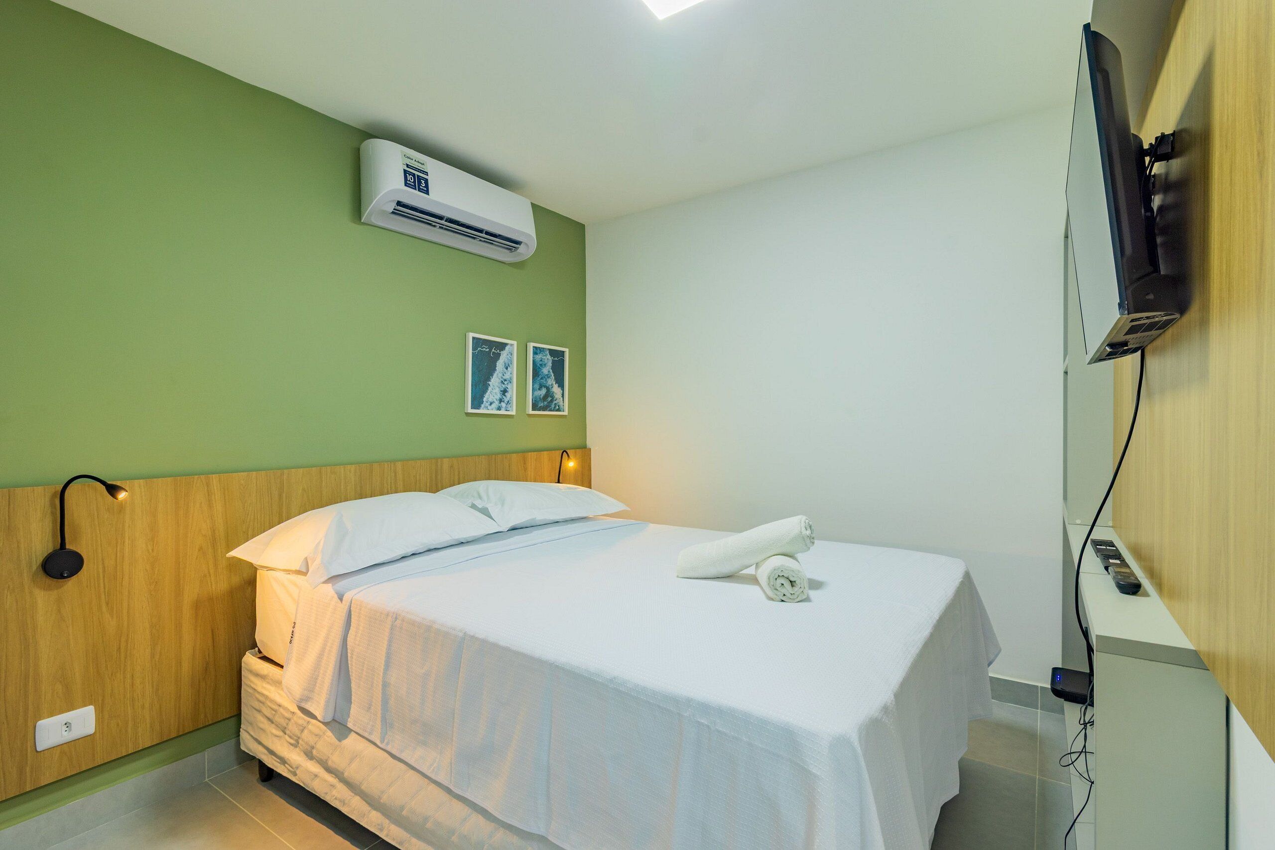 A hotel room with a bed, a wall-mounted air conditioner, a bedside lamp, and a flat-screen TV.