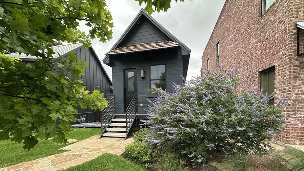 Un bâtiment moderne à l’extérieur gris, avec un escalier menant à l’entrée et un jardin bien entretenu où poussent diverses plantes et fleurs.