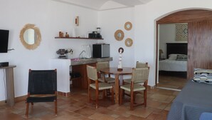 Quarto compacto com mesa de jantar, cadeiras, televisão, cozinha compacta e uma cama no quarto vizinho.