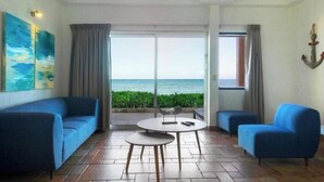 Quarto com uma grande janela, um sofá azul, uma mesa redonda e vista para o oceano.