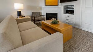 Een woonkamer met een beige bank, een houten salontafel met een decoratief middelpunt, een gestreept fauteuil, een flatscreen televisie boven een open haard, en een witte deur.