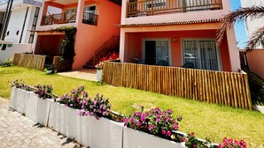 Un edificio residencial de dos pisos con un balcón, cerca viva de madera y un jardín con flores rosas.
