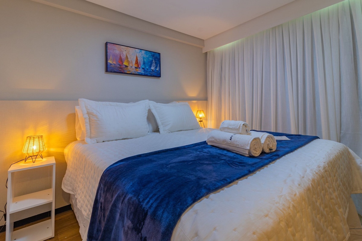 A neatly made bed with white linens and a blue blanket, a painting on the wall, and a white nightstand with a lamp.