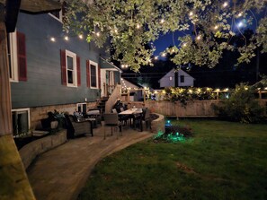 Un patio trasero con terraza, mobiliario exterior e iluminacion con luces colgantes.
