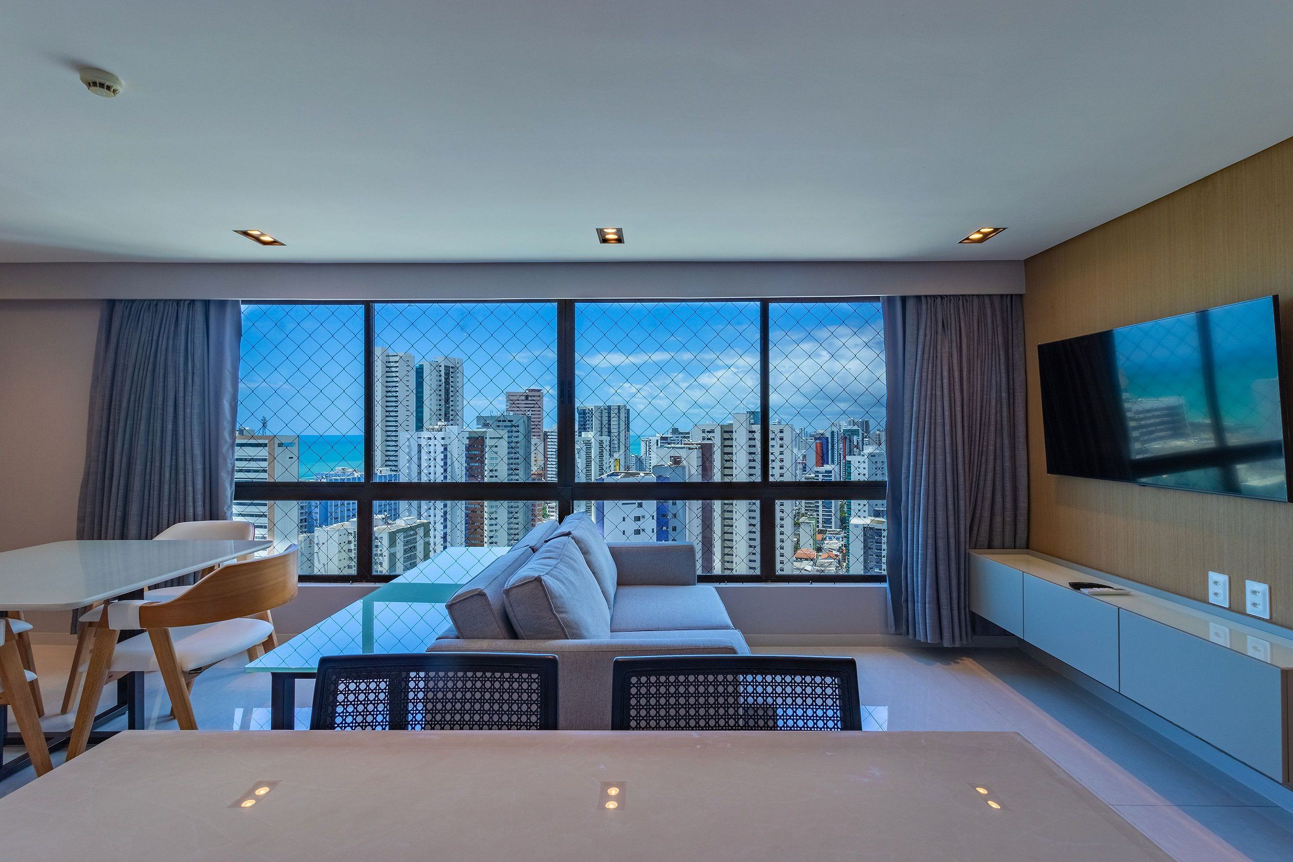 A modern living room with a city view, a sofa, a dining table, and a TV mounted on the wall.