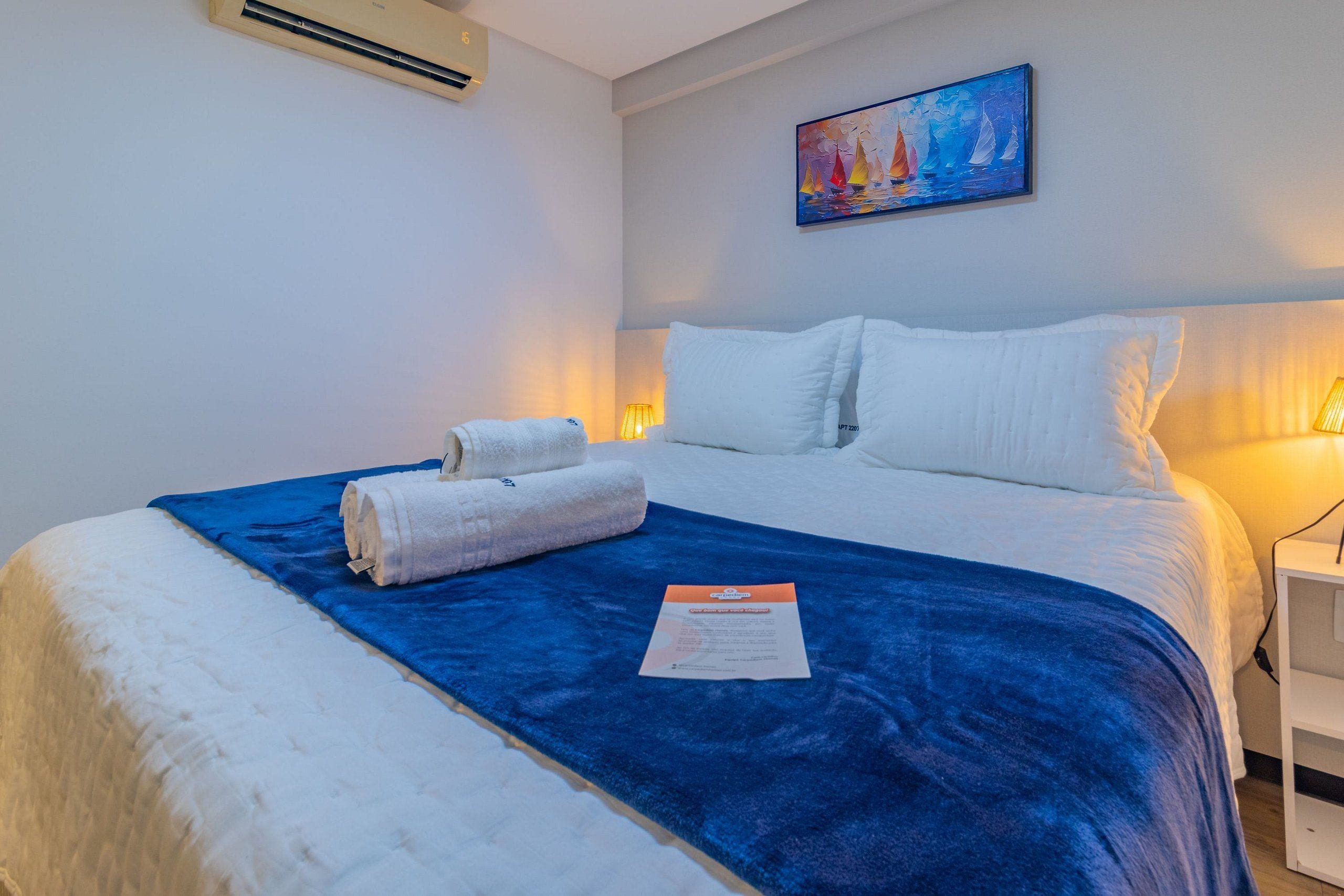 A neatly made bed with white linens and a blue blanket, a painting of sailboats on the wall, and a small bedside table with a lamp.