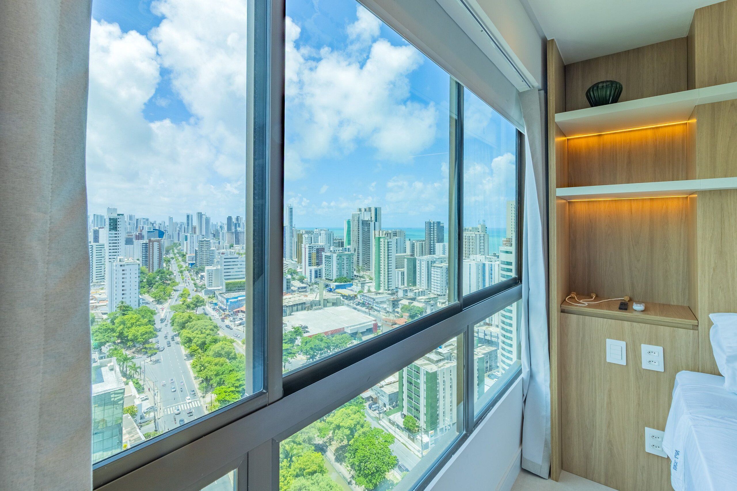 A modern hotel room with large windows offering a city view, a wooden shelf with a bowl, and a bed with a white cover.