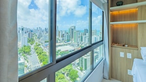 Habitación de hotel moderna con ventanas amplias que ofrecen vistas a la ciudad, un estante de madera con un cuenco y una cama con cubrecama blanco.