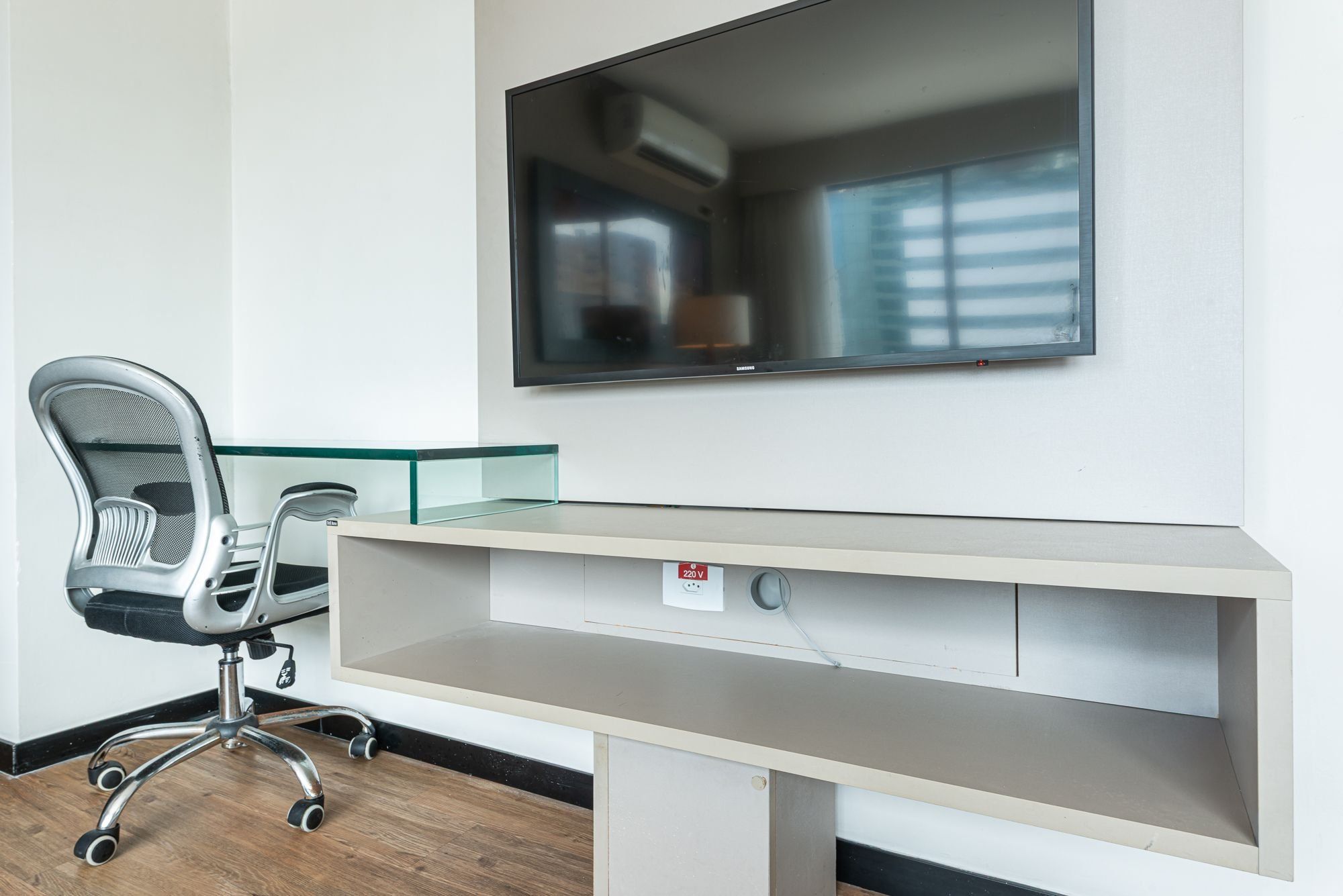 A modern office desk with a glass top, a chair, and a mounted flat-screen TV.