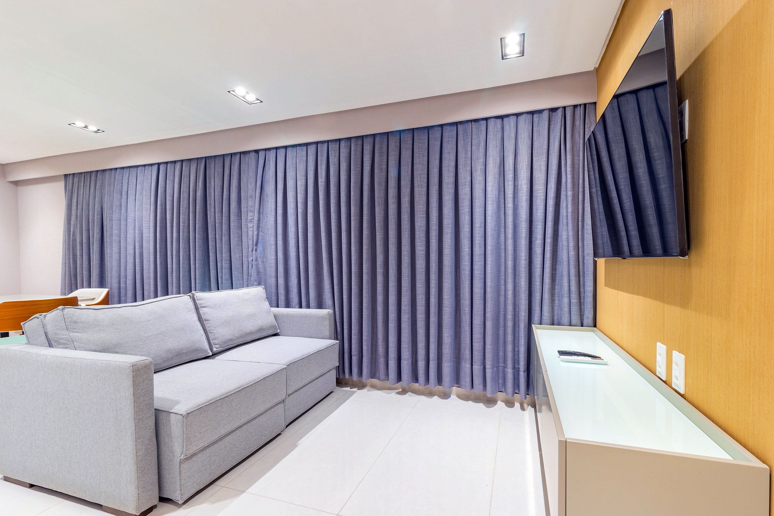 A modern hotel room with a grey sofa, a flat-screen TV mounted on the wall, and a wooden sideboard.
