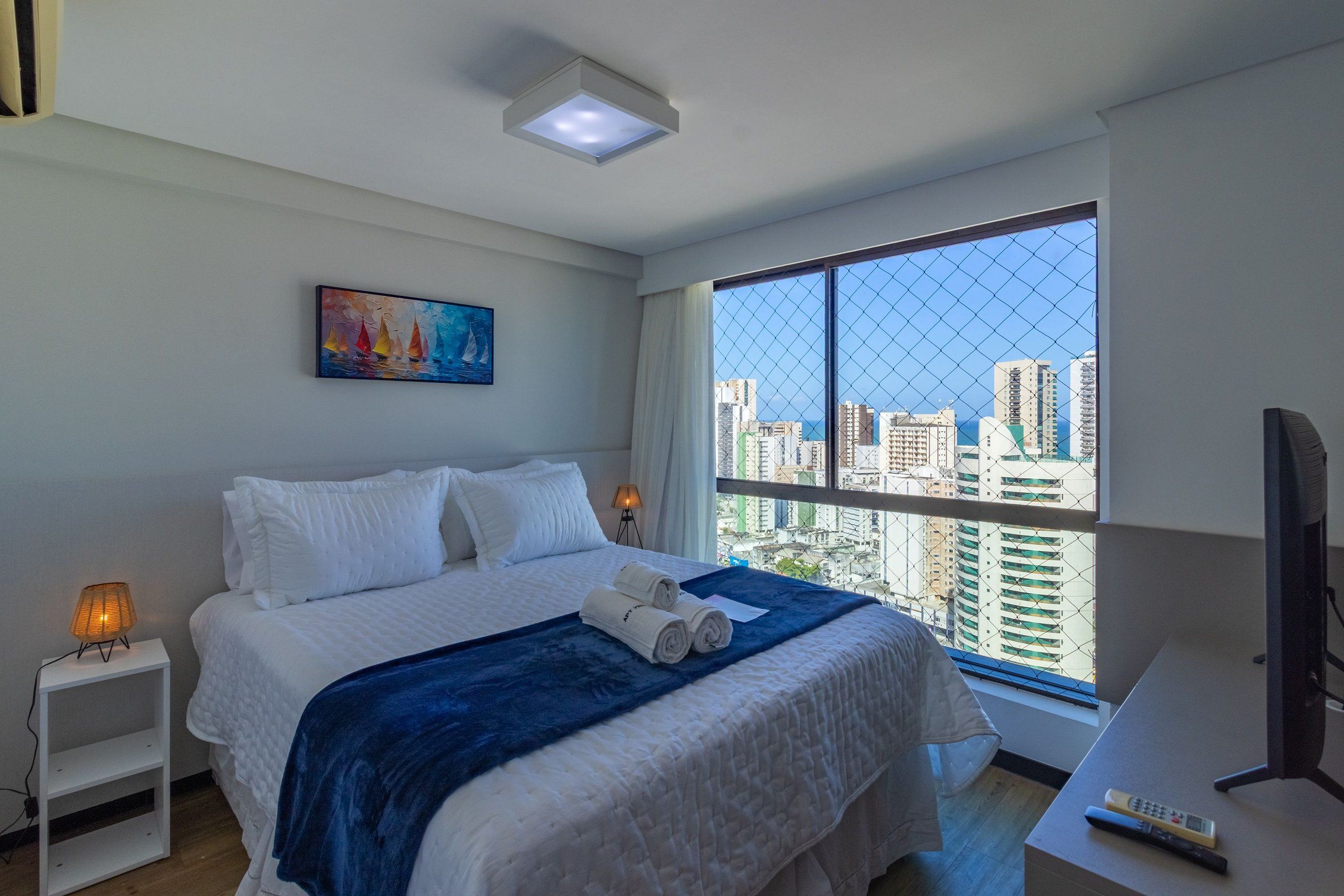 A hotel room with a large window, a bed with white bedding, a painting on the wall, and a desk with a remote control.