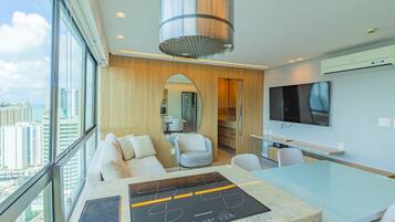 A modern hotel room with a large window, a dining area, a kitchen, and a TV.