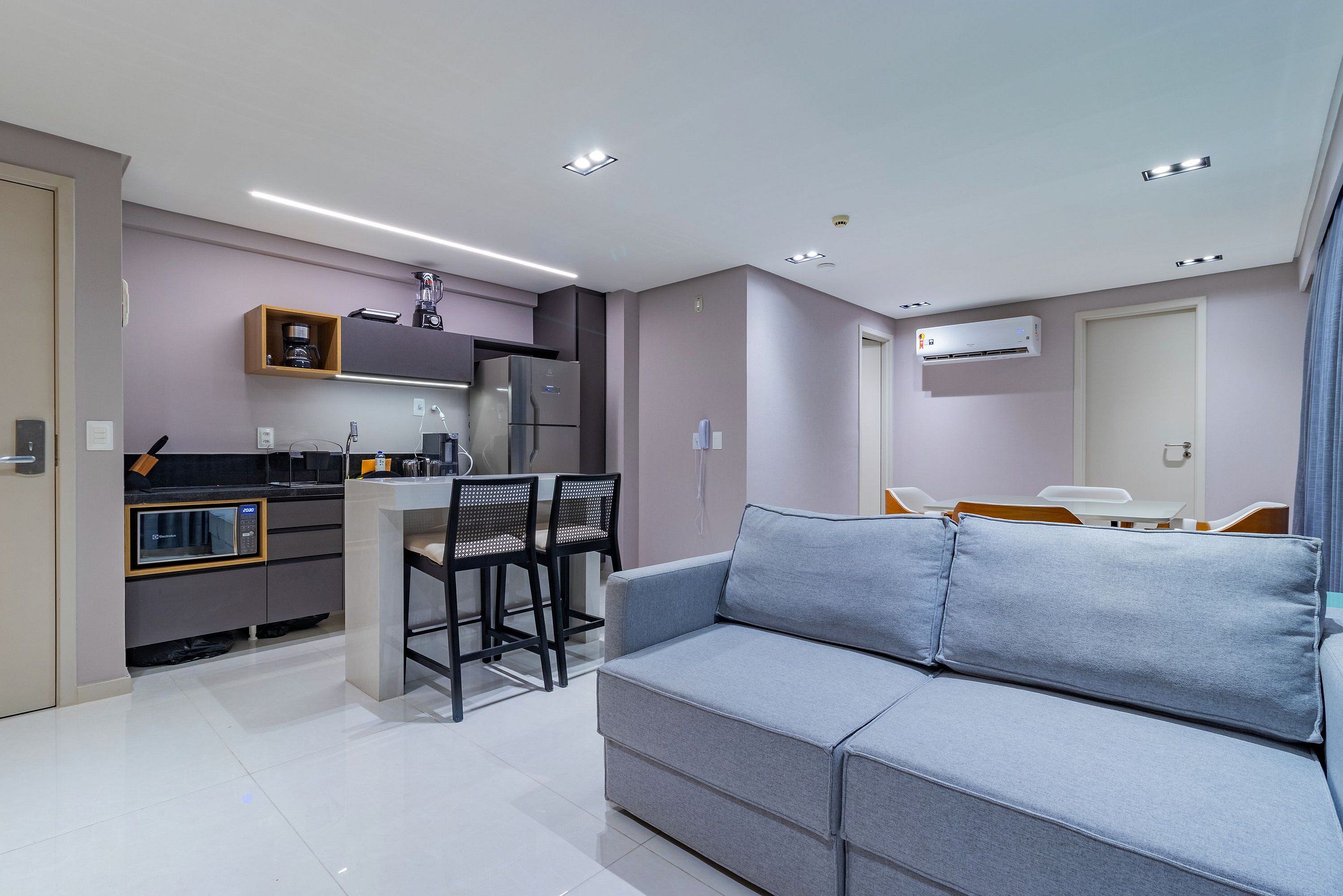 A modern living room with a grey sofa, a dining area with chairs, a kitchen with a refrigerator, and an air conditioning unit.