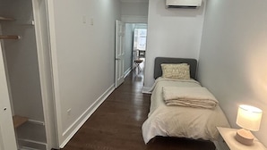 Un couloir étroit, avec un lit au fond, une chaise et un appareil de climatisation fixé au mur.