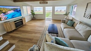 A living room with a TV, wooden flooring, a sofa, and a view of the beach. - Oceanfront house in charming Ewa Beach with AC (Ewa Beach)