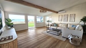 A spacious living room with a large window overlooking the beach, a wooden ceiling, and a modern sofa. - Oceanfront house in charming Ewa Beach with AC (Ewa Beach)