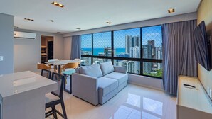 A modern living room with a grey sofa, dining table, and chairs. A large window offers a city view. - Homes Stay #2307 - Flat em Recife by Carpediem (Recife)