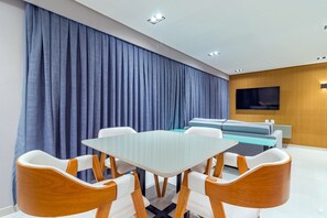 A modern dining area with a glass table and wooden chairs, a sofa, and a TV in the background. - Homes Stay #2307 - Flat em Recife by Carpediem (Recife)