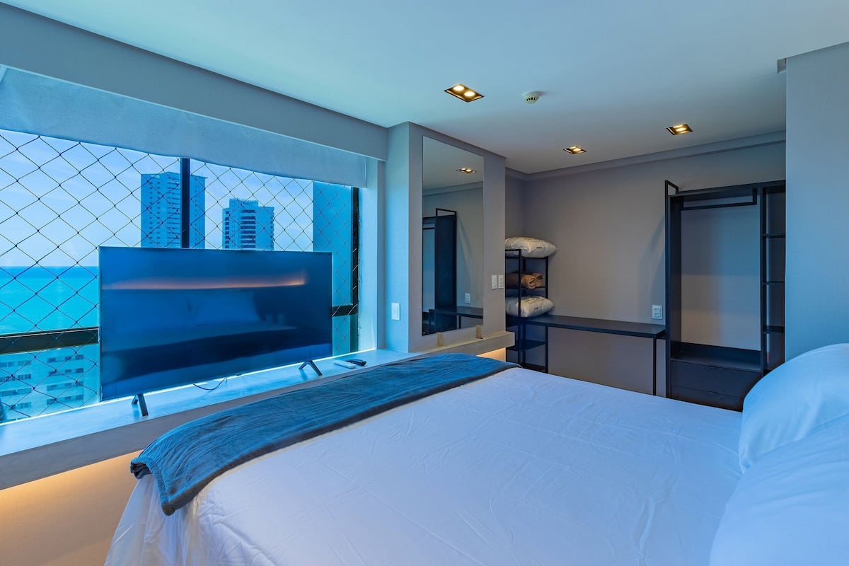 Habitación de hotel moderna con una cama grande, televisión, espejo y vista de la ciudad a través de una ventana.