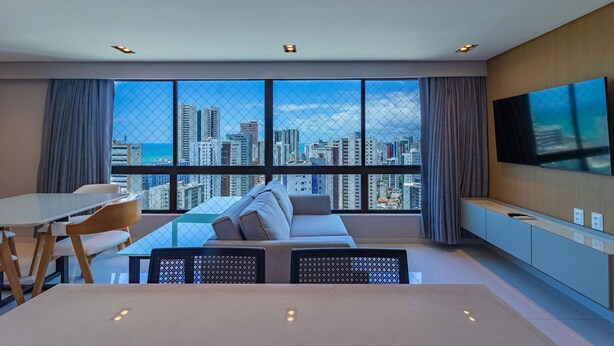 A modern living room with a city view, a sofa, a dining table, and a TV mounted on the wall. - Homes Stay #2307 - Flat em Recife by Carpediem (Recife)