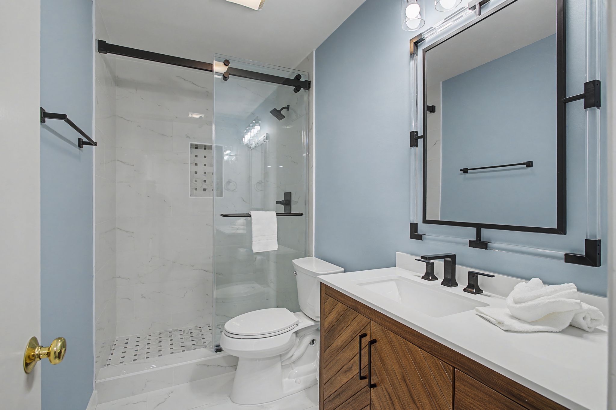 A modern bathroom with a glass-enclosed shower, a white toilet, a wooden vanity with a sink, and a large mirror.