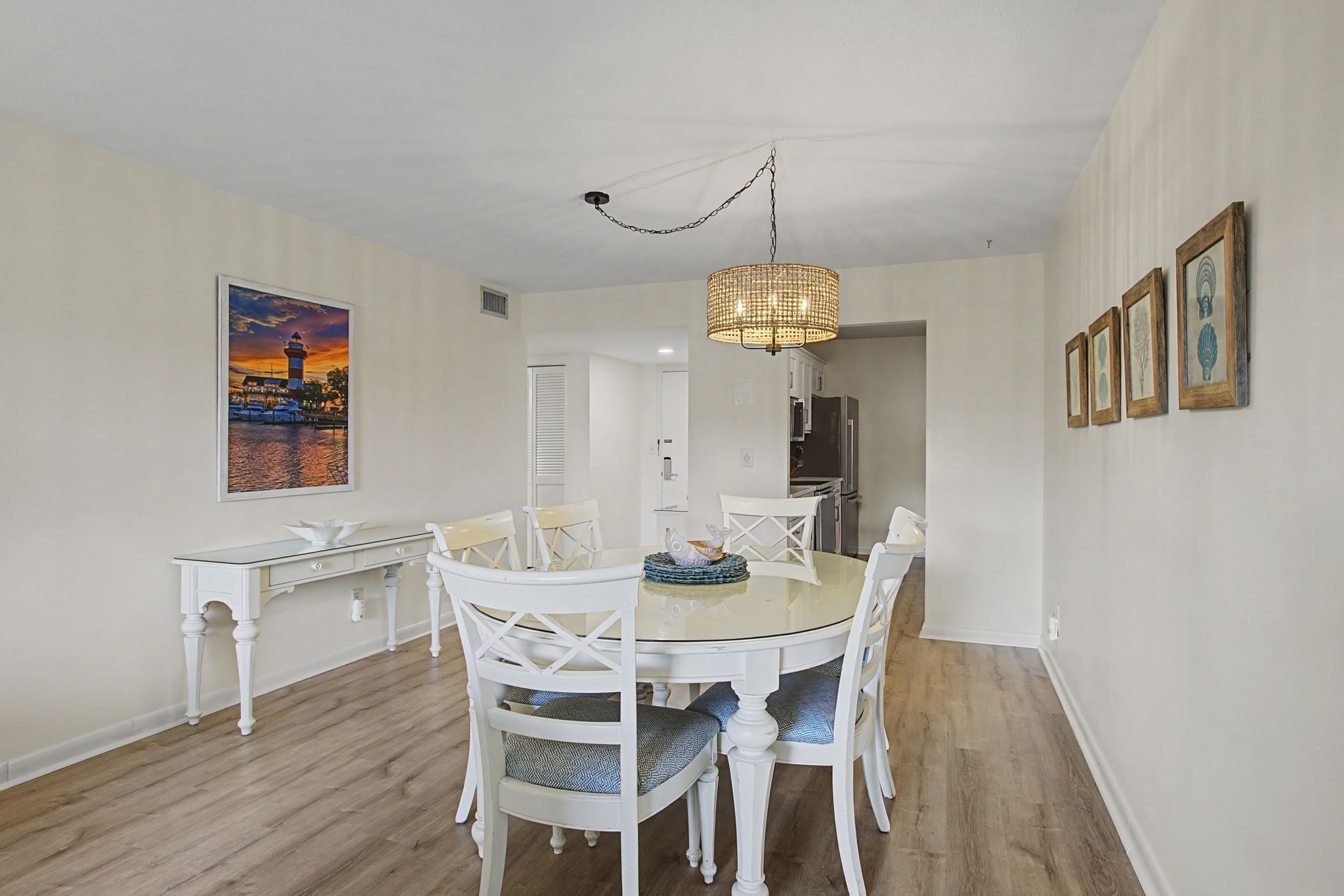 A dining area with a round glass table, white chairs, a hanging light fixture, and a framed painting on the wall.