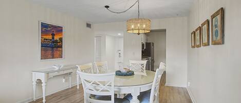 A dining area with a round glass table, white chairs, a hanging light fixture, and a framed painting on the wall.