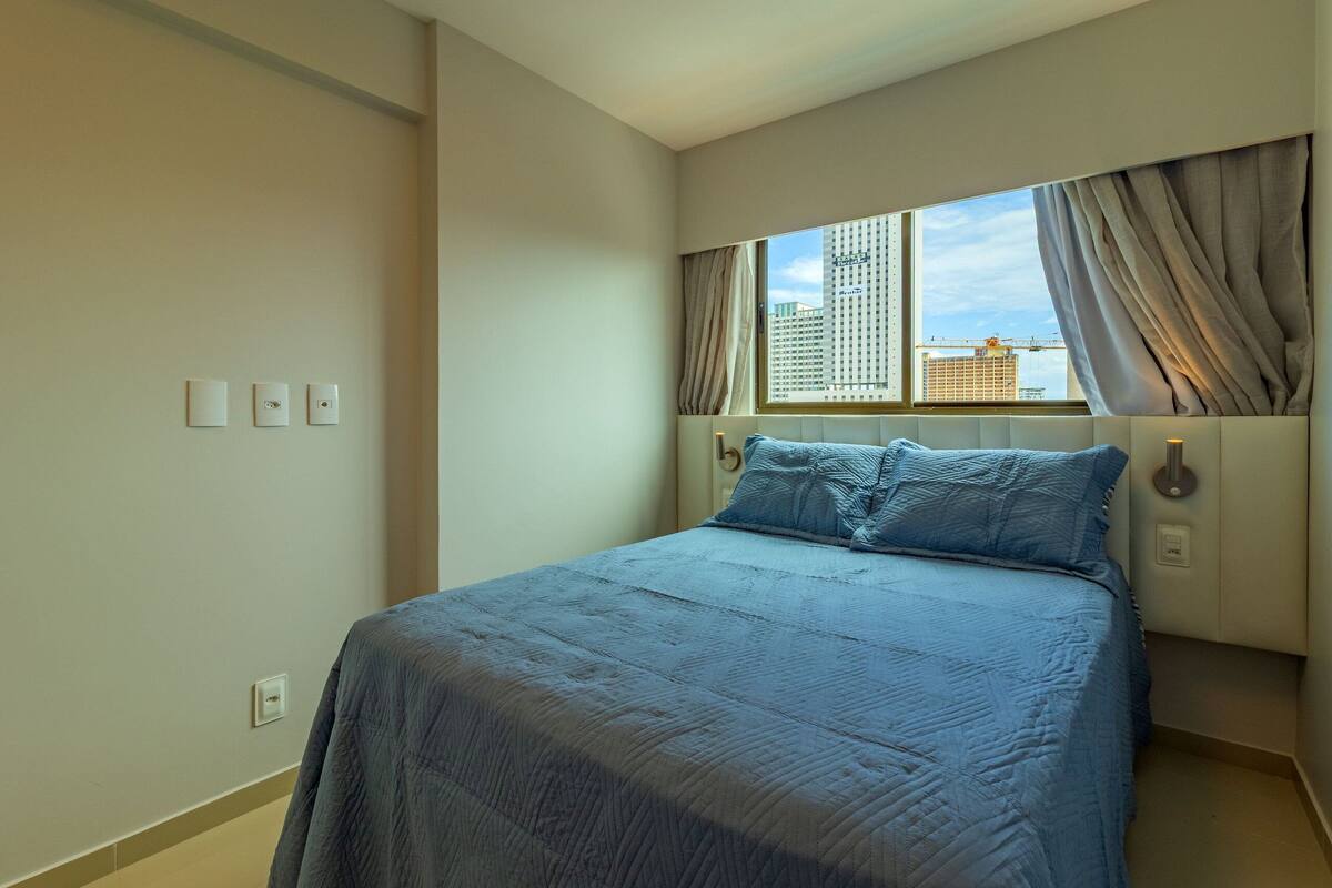 A bedroom with a large bed, a window with curtains, and a view of buildings outside.