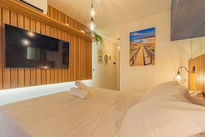A hotel room with a wooden panel wall, a flat-screen TV, a painting of a beach, and a neatly made bed. - Varandas #415 - Porto de Galinhas por Carpediem (Ipojuca)