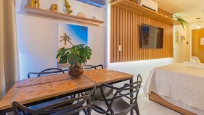 A modern hotel room with a wooden dining table, chairs, a TV mounted on the wall, a bed with a white bedspread, and a potted plant. - Varandas #415 - Porto de Galinhas por Carpediem (Ipojuca)