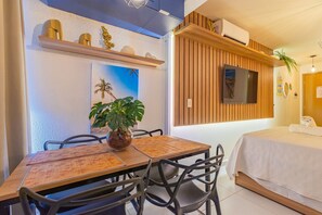 A modern hotel room with a wooden dining table, chairs, a TV mounted on the wall, a bed with a white bedspread, and a potted plant. - Varandas #415 - Porto de Galinhas por Carpediem (Ipojuca)