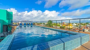 A rooftop swimming pool with clear blue water, surrounded by a glass railing, and overlooking a cityscape with buildings and palm trees. - Varandas #415 - Porto de Galinhas por Carpediem (Ipojuca)