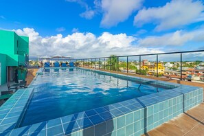 A rooftop swimming pool with clear blue water, surrounded by a glass railing, and overlooking a cityscape with buildings and palm trees. - Varandas #415 - Porto de Galinhas por Carpediem (Ipojuca)