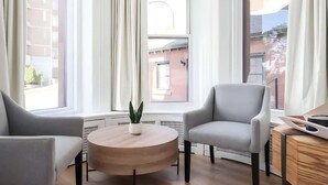 A modern room with two grey armchairs, a wooden coffee table with a plant, and a view of buildings through large windows.