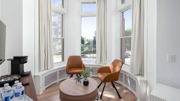Une pièce d’angle avec deux fauteuils marron, une table ronde en bois et une vue sur la rue grâce à de grandes fenêtres.
