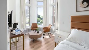Une chambre d’hôtel moderne équipée d’un lit, de deux fauteuils, d’une petite table, d’un téléviseur et de grandes fenêtres donnant sur l’extérieur.