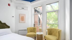 A hotel room with a bed, two yellow armchairs, a small round table with a vase, a large window, and a ceiling light fixture.