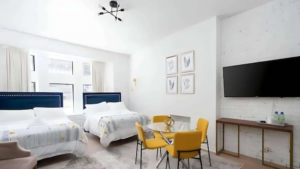 A modern hotel room with two beds, a dining table with yellow chairs, a flat-screen TV, and a white brick wall. - Downtown Gem |2-Beds Condo • Prime location & Wifi (Montreal)