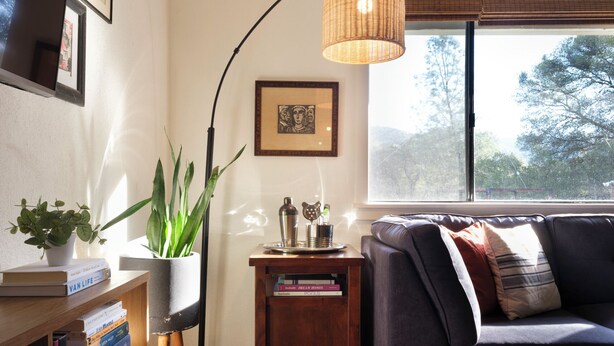 A cozy living room with a dark sofa, a side table with books, a floor lamp, and a large window with a view of trees. - Luxe Yosemite Retreat With Pool + Hot Tub + Game Room (Mariposa)