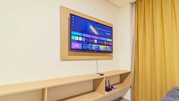 Ein Hotelzimmer mit einem an der Wand befestigten Flachbildfernseher, der einen Startbildschirm zeigt, einem Holztischchen und einem gelben Vorhang.