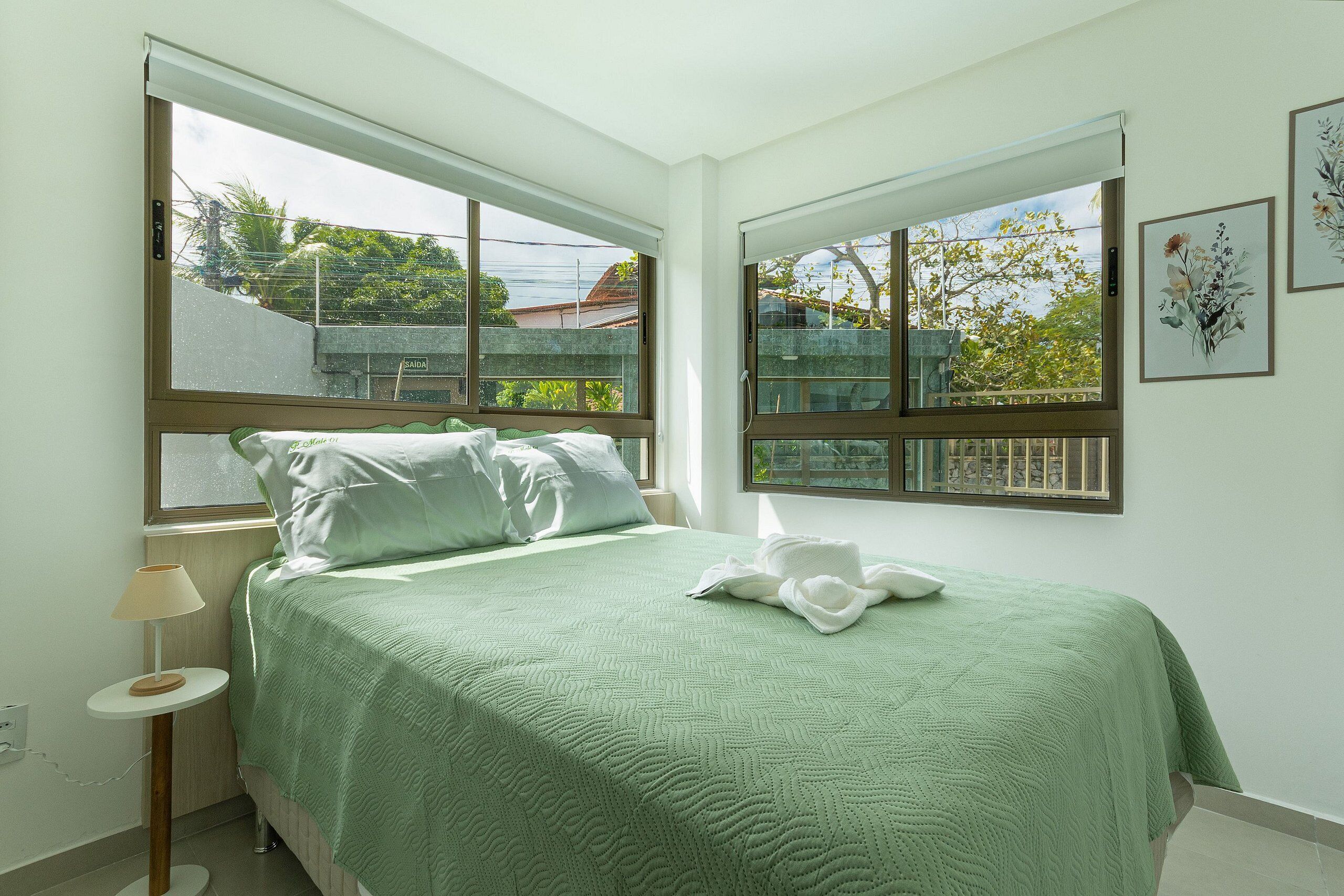 A bedroom with a bed, a bedside table, a lamp, and two windows with views of greenery and buildings.