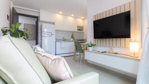 Een moderne woonkamer met een bank, een eethoek met tafel en stoelen, een keuken met witte kasten, een koelkast en een flatscreen televisie aan de muur.