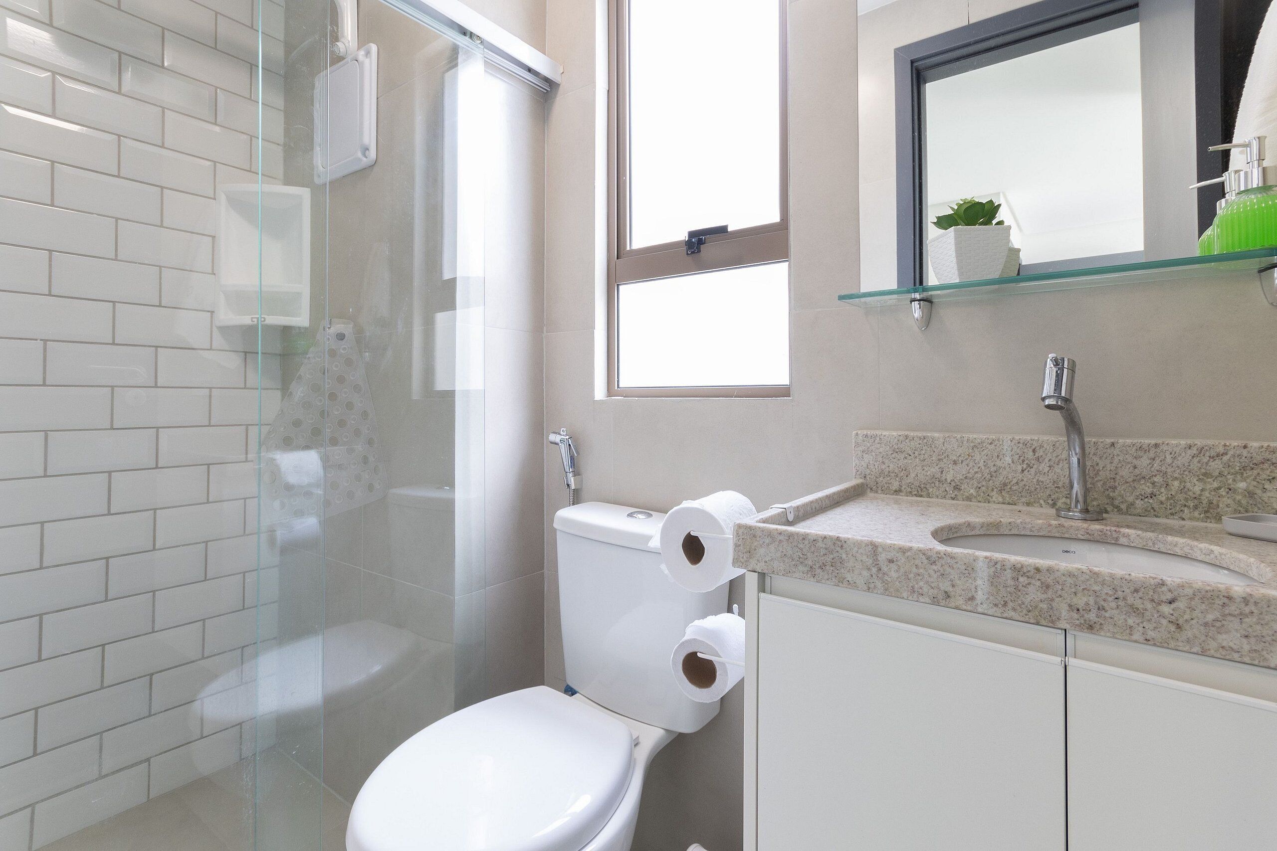 A modern bathroom with a glass shower enclosure, a toilet with two rolls of toilet paper, a sink with a granite countertop, and a mirror above the sink.