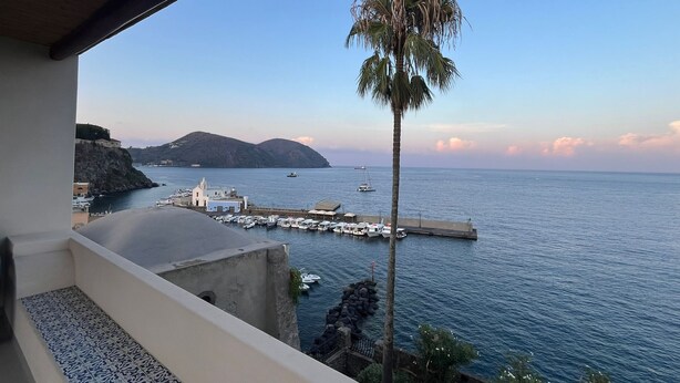 Vista costera desde un balcón con una palmera, un muelle, barcos y una montaña al fondo.
