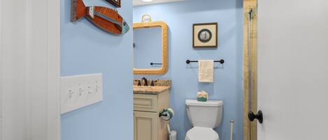 A bathroom with a white toilet, a sink with a marble countertop, a towel rack with a white towel, a framed mirror, and a framed picture on the wall.