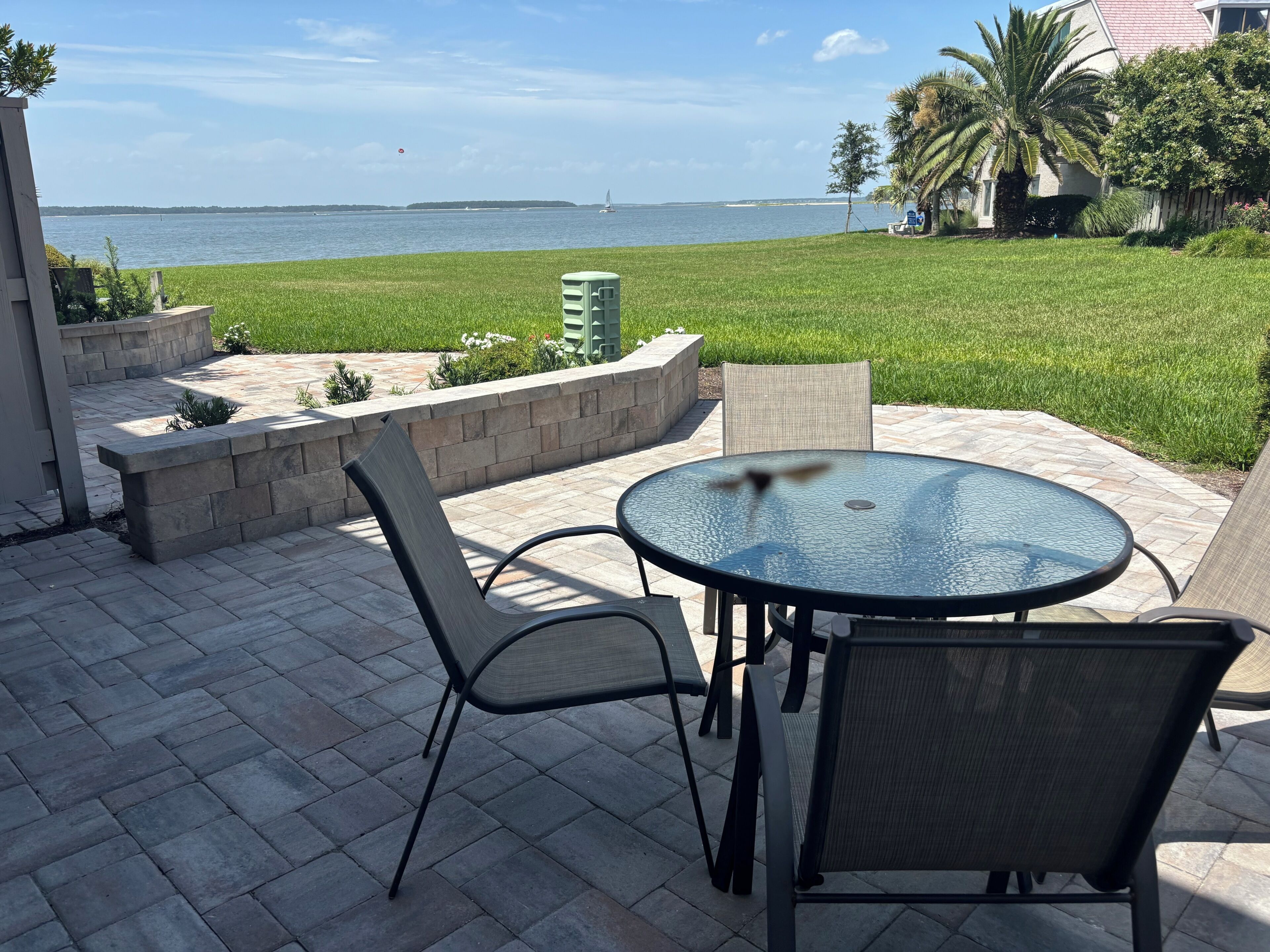 An outdoor patio with a table and chairs, a view of the sea, and a grassy area with a palm tree.