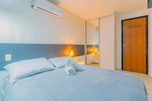 A neatly made bed with white linens and a rolled towel, a bedside lamp, and a wooden door with a small mirror. - Porto Plaza #210 - Triple Flat by Carpediem (Ipojuca)
